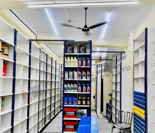 Slotted angle racks , blue and white rack with boxes on the first 2 shelves and rest of the shelves contain moror oil of different kinds and company. the shop/warehouse has white interior and white shelves with white light and a brown fan. Only the angles are blue and the plastic crates used for storing small metal clunks like nuts and bolts are blue in color.