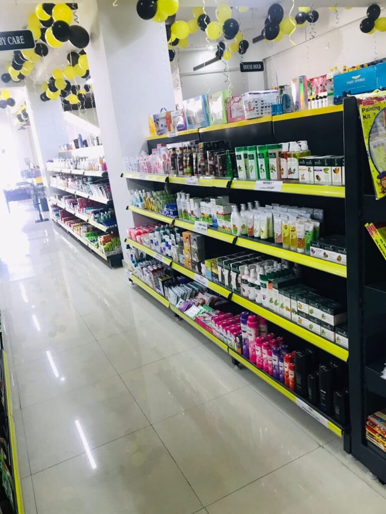 Display Racks for Shop in Dehradun Shelving: Multiple rows of black shelves with bright yellow edges, stocked with various food products. Products: A wide variety of cereals, including recognizable brands like Kellogg's Many types of snacks, particularly corn-based snacks like tortilla chips Different flavors and brands of chips and crunchy snacks Some oatmeal and breakfast products Organization: The products are neatly arranged and grouped by type and brand. Signage: There's a small sign visible that says "CEREALS" indicating the product category. Decoration: The ceiling is decorated with balloons in black and yellow colors, along with some streamers. This suggests the store may be celebrating an opening, anniversary, or special event. Lighting: Bright fluorescent lights hang from the ceiling, providing ample illumination for the products. Layout: The shelves are arranged in an L-shape, with the corner of the aisle visible in the image.