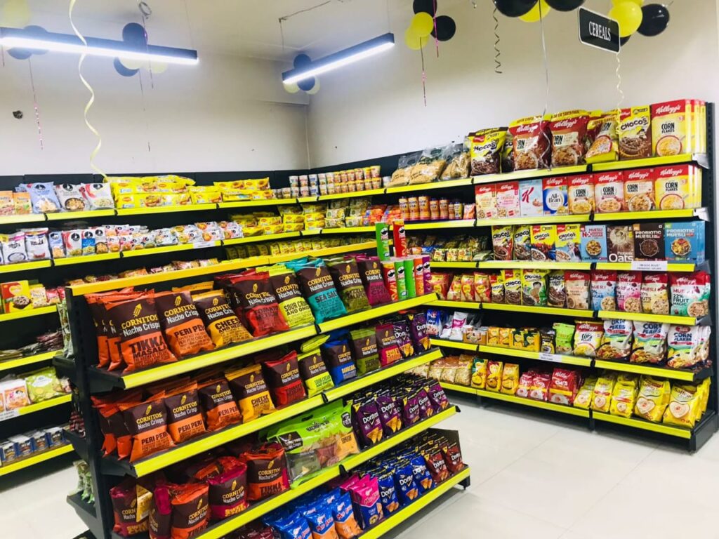 Display Racks for Shop in Dehradun Shelving: Multiple rows of black shelves with bright yellow edges, stocked with various food products. Products: A wide variety of cereals, including recognizable brands like Kellogg's Many types of snacks, particularly corn-based snacks like tortilla chips Different flavors and brands of chips and crunchy snacks Some oatmeal and breakfast products Organization: The products are neatly arranged and grouped by type and brand. Signage: There's a small sign visible that says "CEREALS" indicating the product category. Decoration: The ceiling is decorated with balloons in black and yellow colors, along with some streamers. This suggests the store may be celebrating an opening, anniversary, or special event. Lighting: Bright fluorescent lights hang from the ceiling, providing ample illumination for the products. Layout: The shelves are arranged in an L-shape, with the corner of the aisle visible in the image.