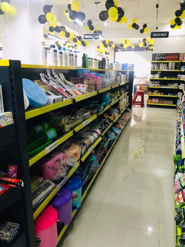 Display Racks for Shop in Dehradun Shelving: Multiple rows of black shelves with bright yellow edges, stocked with various food products. Products: A wide variety of cereals, including recognizable brands like Kellogg's Many types of snacks, particularly corn-based snacks like tortilla chips Different flavors and brands of chips and crunchy snacks Some oatmeal and breakfast products Organization: The products are neatly arranged and grouped by type and brand. Signage: There's a small sign visible that says "CEREALS" indicating the product category. Decoration: The ceiling is decorated with balloons in black and yellow colors, along with some streamers. This suggests the store may be celebrating an opening, anniversary, or special event. Lighting: Bright fluorescent lights hang from the ceiling, providing ample illumination for the products. Layout: The shelves are arranged in an L-shape, with the corner of the aisle visible in the image.