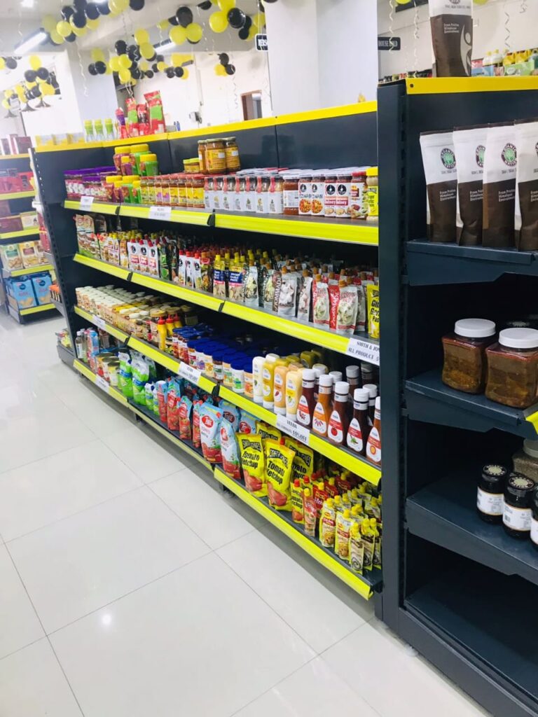 Display Racks for Shop in Dehradun Shelving: Multiple rows of black shelves with bright yellow edges, stocked with various food products. Products: A wide variety of cereals, including recognizable brands like Kellogg's Many types of snacks, particularly corn-based snacks like tortilla chips Different flavors and brands of chips and crunchy snacks Some oatmeal and breakfast products Organization: The products are neatly arranged and grouped by type and brand. Signage: There's a small sign visible that says "CEREALS" indicating the product category. Decoration: The ceiling is decorated with balloons in black and yellow colors, along with some streamers. This suggests the store may be celebrating an opening, anniversary, or special event. Lighting: Bright fluorescent lights hang from the ceiling, providing ample illumination for the products. Layout: The shelves are arranged in an L-shape, with the corner of the aisle visible in the image.