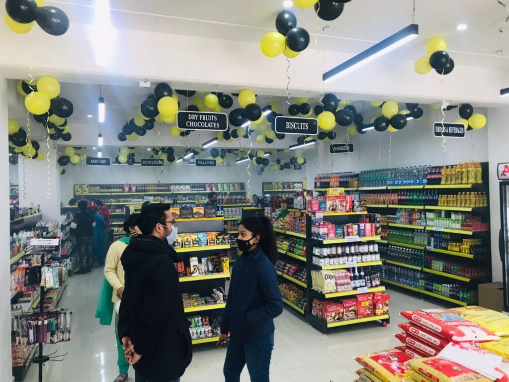 Display Racks for Shop in Dehradun Shelving: Multiple rows of black shelves with bright yellow edges, stocked with various food products. Products: A wide variety of cereals, including recognizable brands like Kellogg's Many types of snacks, particularly corn-based snacks like tortilla chips Different flavors and brands of chips and crunchy snacks Some oatmeal and breakfast products Organization: The products are neatly arranged and grouped by type and brand. Signage: There's a small sign visible that says "CEREALS" indicating the product category. Decoration: The ceiling is decorated with balloons in black and yellow colors, along with some streamers. This suggests the store may be celebrating an opening, anniversary, or special event. Lighting: Bright fluorescent lights hang from the ceiling, providing ample illumination for the products. Layout: The shelves are arranged in an L-shape, with the corner of the aisle visible in the image.